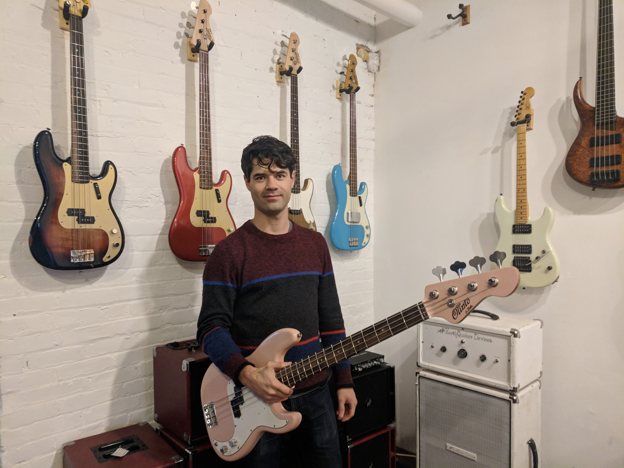 Dan seated in his Denver shop holding an electric bass with instruments on the wall